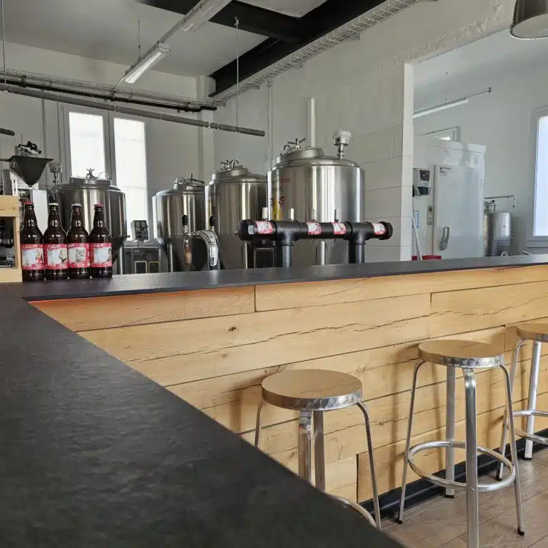 Intérieur d'une brasserie artisanale avec des cuves en acier inoxydable et un bar en bois. Des bouteilles de bière sont alignées sur le comptoir, et des tabourets en métal sont visibles. L'éclairage moderne complète l'ambiance industrielle de l'espace.