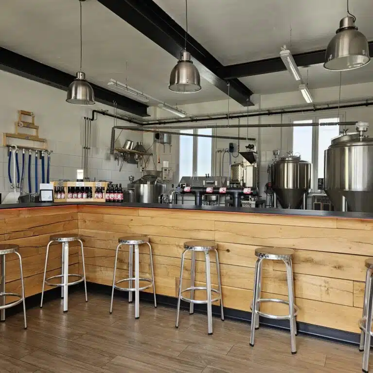 Intérieur d'une brasserie moderne avec un bar en bois, des tabourets en métal, des cuves de fermentation en acier inoxydable et des bouteilles de bière exposées sur le comptoir. Éclairage industriel avec des lampes suspendues et un espace de travail bien équipé.