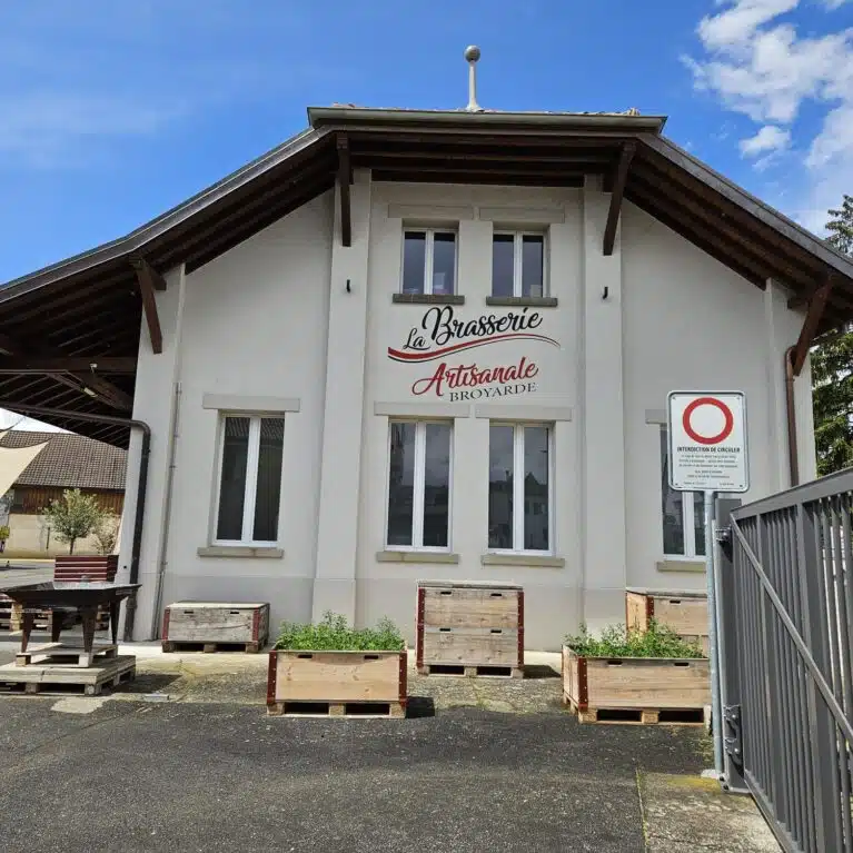 Brasserie artisanale Broyarde avec une façade blanche, des fenêtres modernes, et des jardinières en bois. Le bâtiment est entouré de panneaux de signalisation et d'un auvent en toile, sous un ciel bleu.