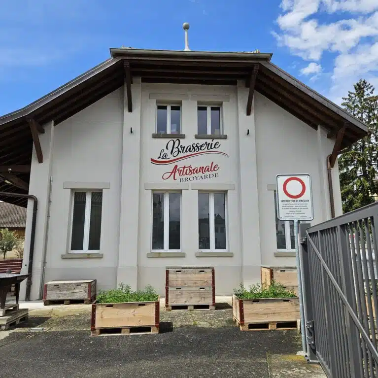 Façade de la brasserie artisanale Broyarde, avec un panneau indiquant le nom et des bacs en bois plantés d'herbes devant l'entrée, sous un ciel bleu parsemé de nuages.