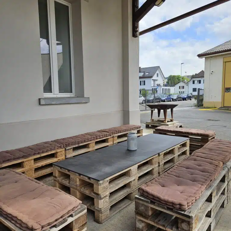 Mobilier extérieur en palettes en bois avec coussins, créant un espace de détente moderne. Une table noire en bois est au centre, entourée de bancs confortables. L'environnement est lumineux avec un ciel partiellement nuageux et des bâtiments en arrière-plan.