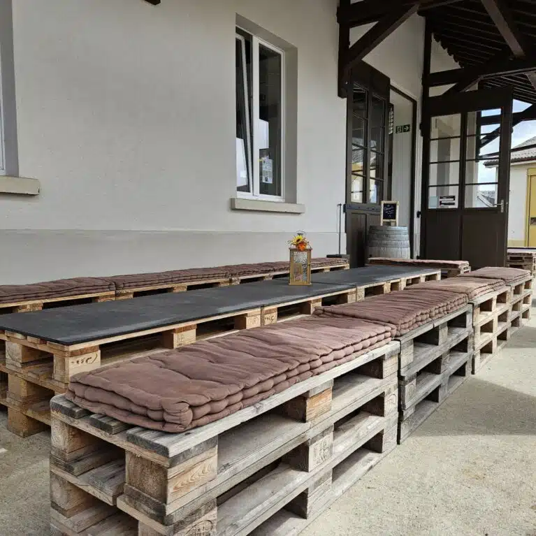 Espace extérieur aménagé avec des bancs en palettes en bois et des coussins, idéal pour se détendre. La table centrale en bois est entourée de palettes, créant une ambiance conviviale près d'une entrée.