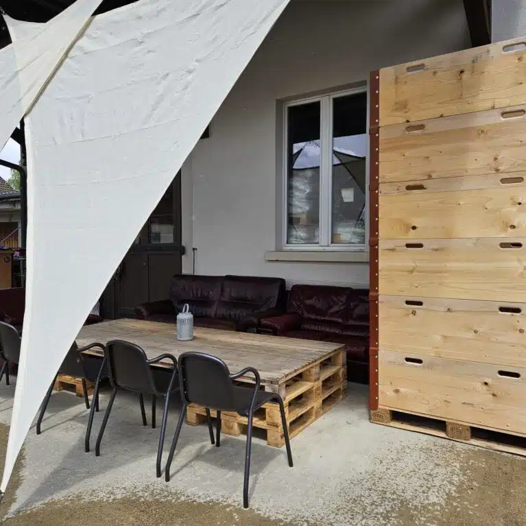 Terrasse extérieure avec une table en palettes, chaises noires et un auvent blanc, offrant un espace de détente confortable près d'une maison.