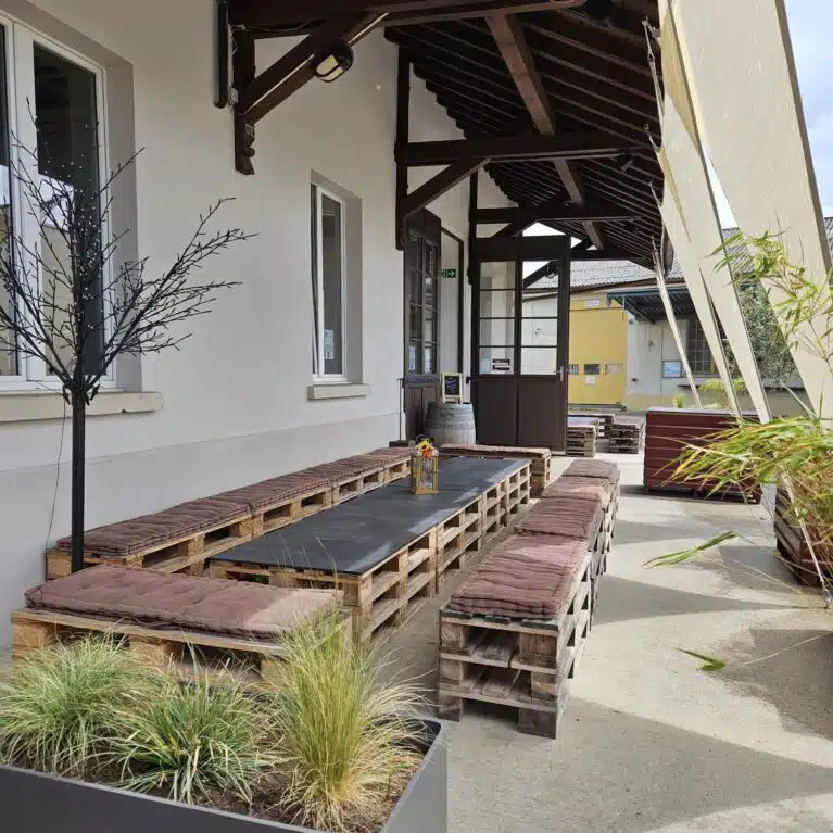 Espace extérieur moderne avec des bancs en palettes, un plateau noir, et des plantes décoratives, sous une structure en bois. Parfait pour se détendre ou socialiser.